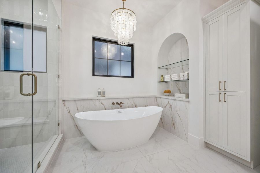 Another view of the 71" Soaking Tub with a Wall-Mounted Tub Filler in Champagne Bronze and a Light Glossy Bronze Finish Tiered Chandelier with Natural White Shell.   There is also open glass shelving in the alcove wall that holds neatly stacked towels, jars, and bath accessories.  Linen Storage Closet for Toiletries and Towels.