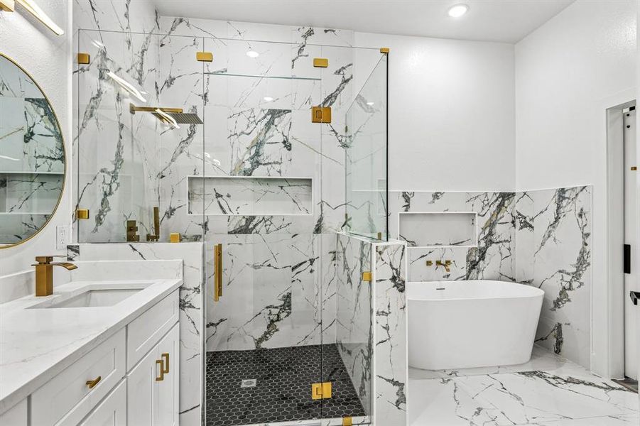 Luxurious bathroom featuring a spacious walk-in shower with black hex tile flooring and gold-toned fixtures, a freestanding soaking tub, an integrated vanity sink with gold-toned faucet, a large round mirror, and a full-height tile surround with built-in niches