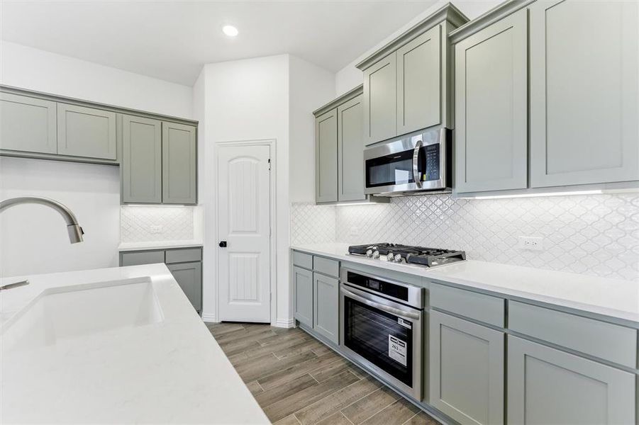 Kitchen with gray cabinetry, light stone counters, wood tiled floors, appliances with stainless steel finishes, and decorative backsplash