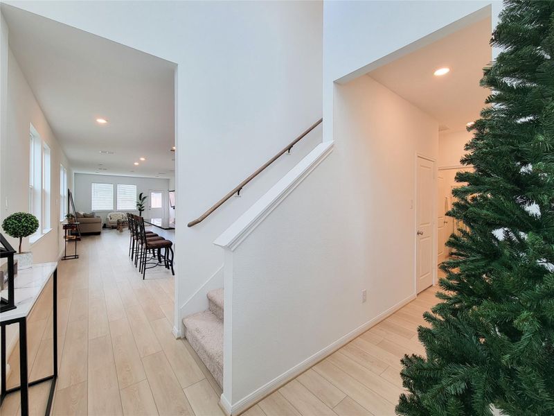 Open entryway with light wood flooring and a staircase leading up. The space flows into a well-lit living area with large windows and a dining section, providing an inviting atmosphere.