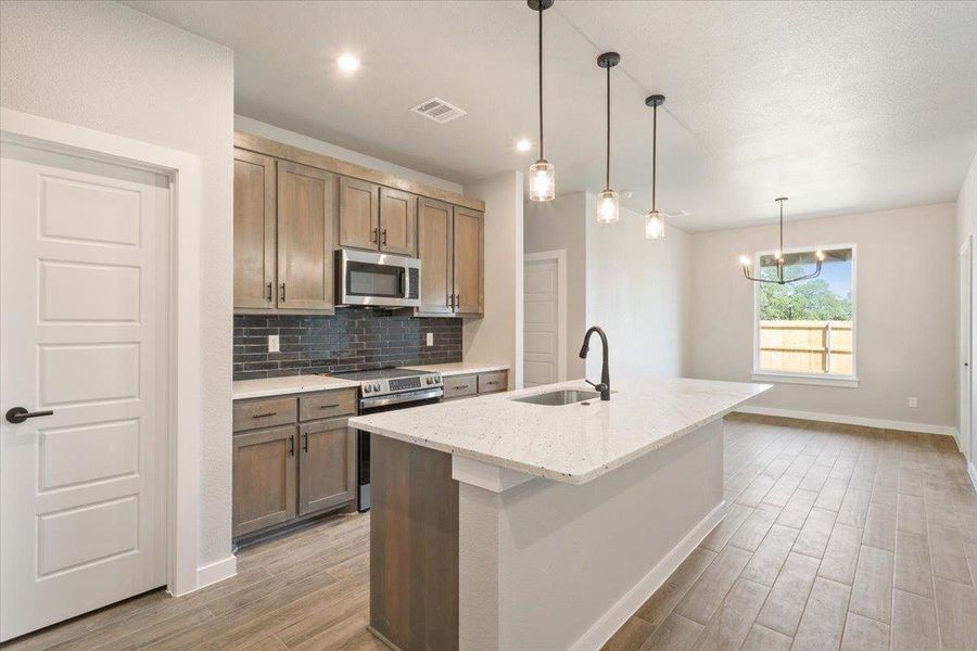 Kitchen featuring backsplash, decorative light fixtures, stainless steel appliances, a kitchen island with sink, and light stone countertops