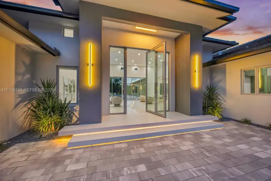 Modern exterior entrance featuring a glass-panel pivoting entry door, dual elevated concrete steps with integrated lighting, sleek vertical wall sconces, and stone paver driveway