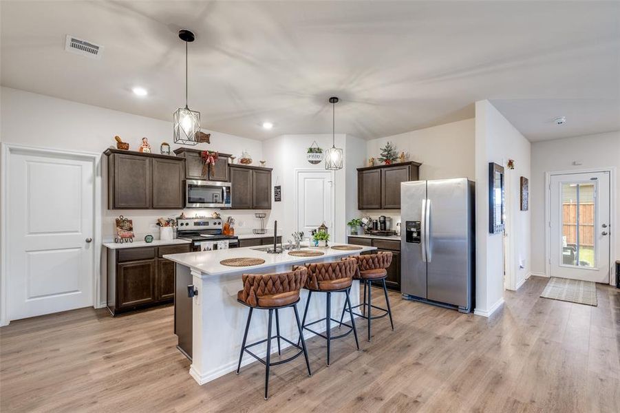 Kitchen with dark brown cabinetry, a breakfast bar area, stainless steel appliances, pendant lighting, and a kitchen island with sink Kitchen with dark brown cabinetry, a breakfast bar area, stainless steel appliances, pendant lighting, and a kitchen island with sink