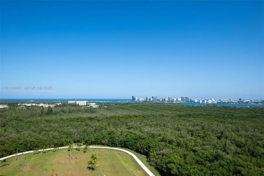 Aerial perspective showcasing extensive natural greenery, open lawn space, paved pathways, coastal water frontage, and a distant cityscape