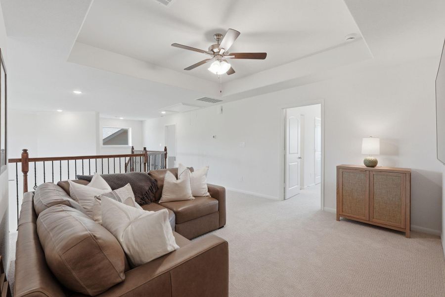 This spacious, well-lit living area features a comfortable sectional sofa, neutral carpeting, and a ceiling fan. It opens to other rooms and has a stylish railing, creating an inviting atmosphere for relaxation or entertaining.