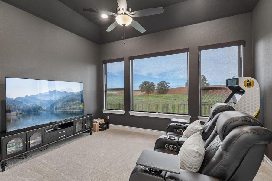 Media room featuring three large windows with roller shades, a ceiling fan with integrated lighting, and neutral-toned carpeting