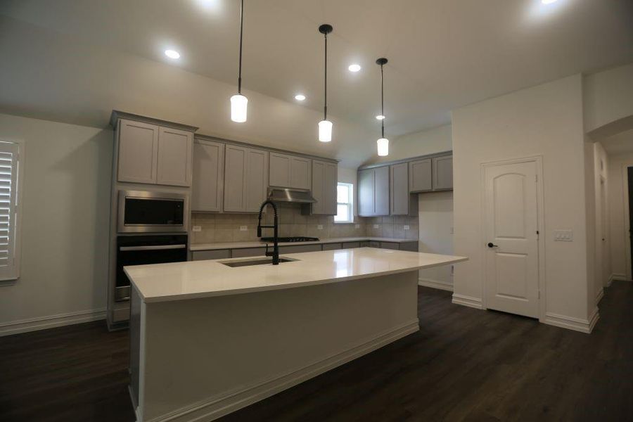 Modern kitchen featuring a large central island with a sink, light gray cabinetry, stainless steel appliances, dark wood-finish flooring, and recessed and pendant lighting
