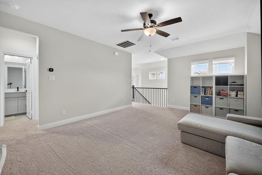 Carpeted room featuring a ceiling fan with light fixture, multiple windows, a baluster railing, and an open doorway revealing a bathroom with a vanity and mirror