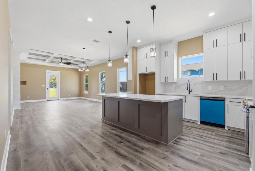 Open-concept kitchen seamlessly flows into the living area and showcases sleek, modern white cabinetry, a custom vent hood, and stunning quartz countertops that extend as a full-height backsplash. It also features a large undermount sink positioned beneath a window for natural light, and an expansive island with three pendant lights.