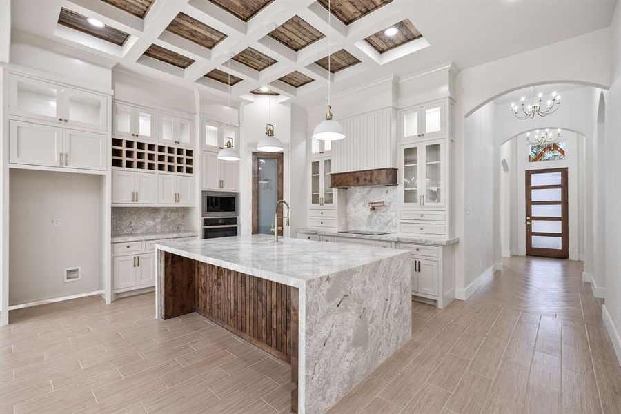 Kitchen with glass insert cabinets, decorative backsplash, coffered ceiling, a kitchen island with sink, and light stone counters