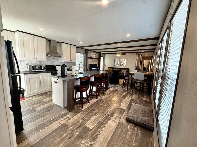Kitchen with dark countertops, a breakfast bar, white cabinetry, a kitchen island with sink, and wall chimney exhaust hood