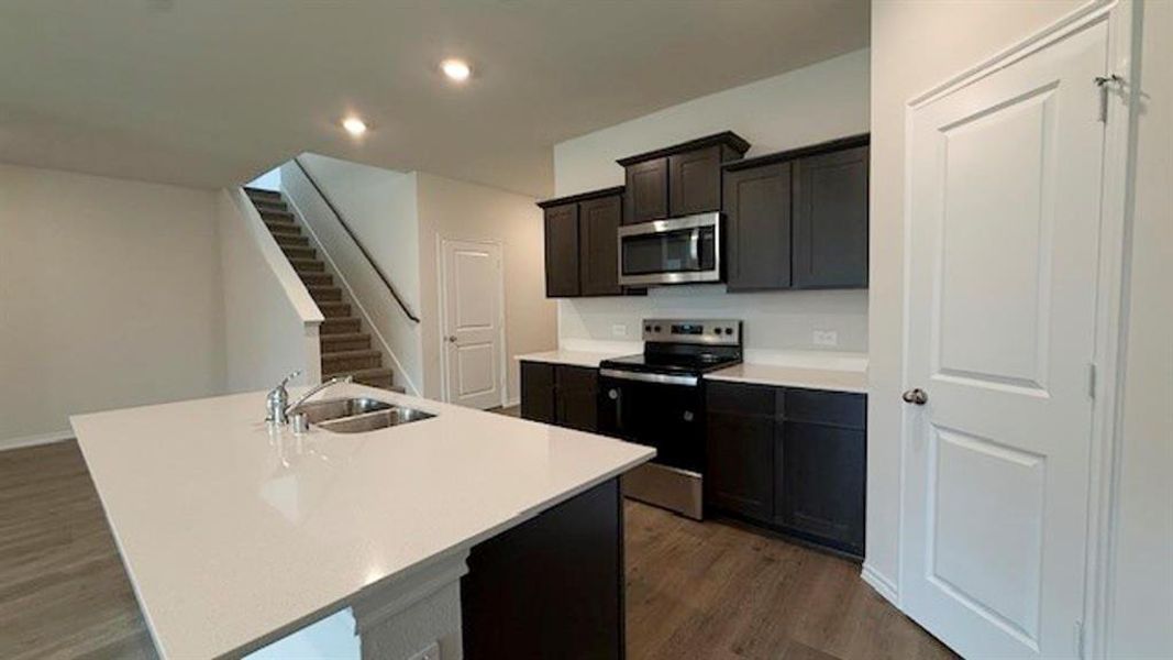 Kitchen with dark wood-type flooring, appliances with stainless steel finishes, an island with sink, recessed lighting, and light stone counters