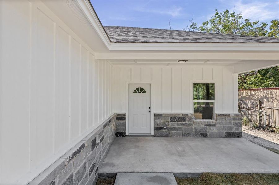 Doorway to property featuring covered porch, board and batten siding, roof with shingles, and stone siding