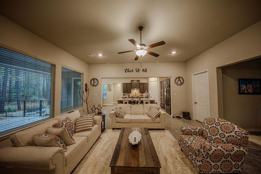 This cozy living room features neutral tones with stylish patterned furniture and a plush rug. Large windows offer plenty of natural light, and an open layout connects to the modern kitchen. Ceiling fan and tasteful decor provide a warm, inviting atmosphere.