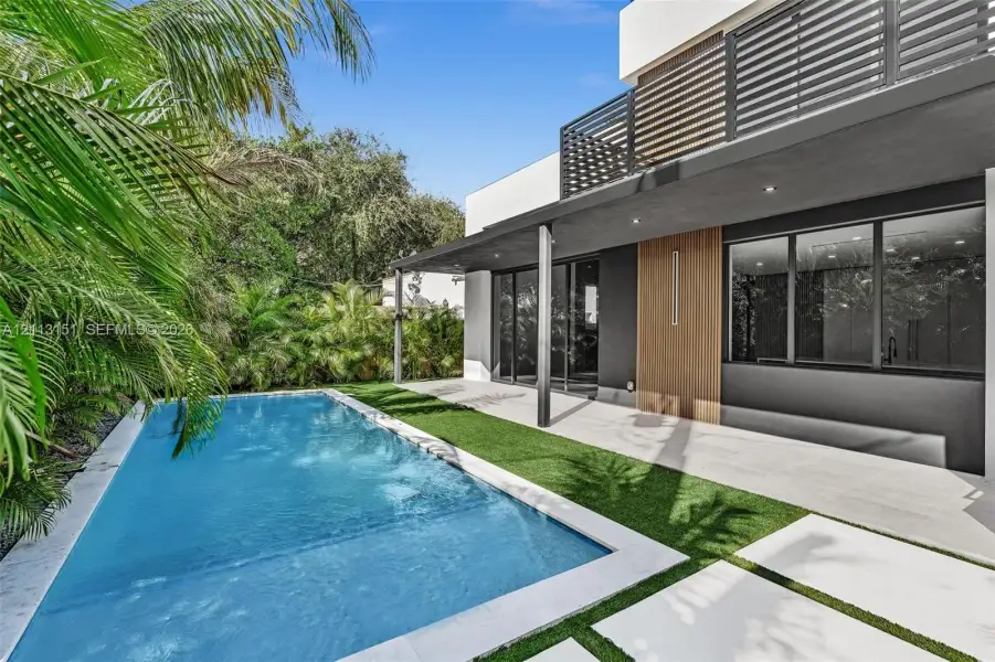 Modern backyard featuring a swimming pool, covered concrete patio, synthetic turf lawn, wood-tone panel siding, and a metal balcony railing Modern backyard featuring a swimming pool, covered concrete patio, synthetic turf lawn, wood-tone panel siding, and a metal balcony railing