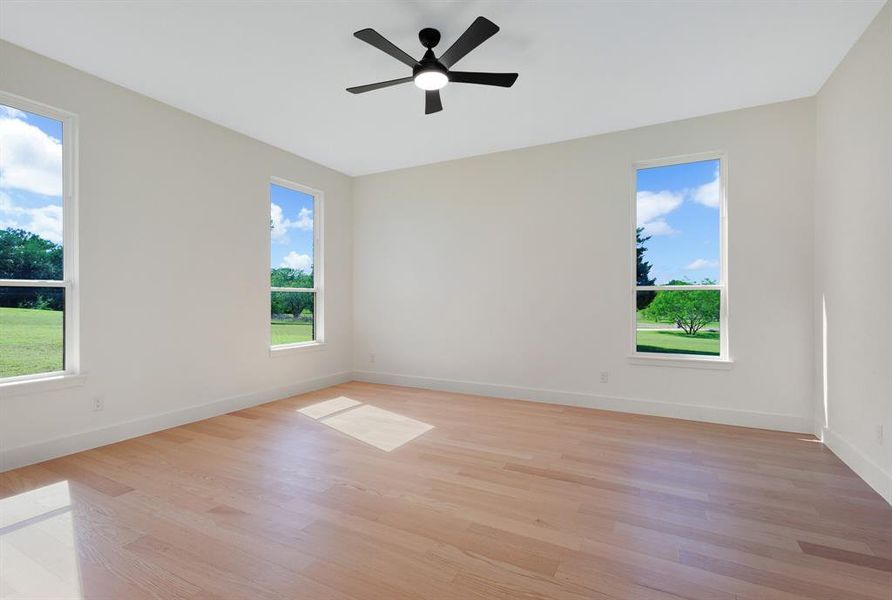 Spacious room featuring light wood-finish flooring, three vertical windows, white baseboards, and a black ceiling fan with integrated lighting Spacious room featuring light wood-finish flooring, three vertical windows, white baseboards, and a black ceiling fan with integrated lighting