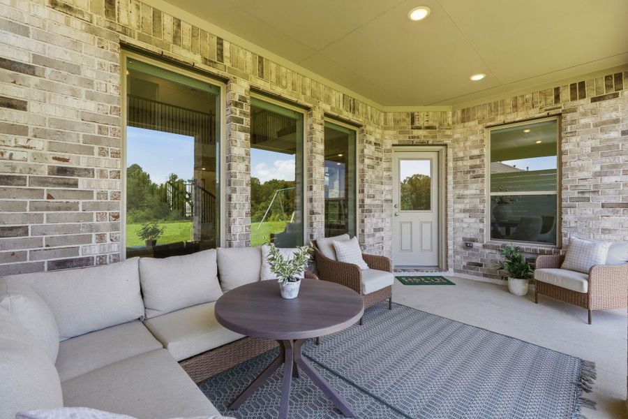 This inviting patio features a cozy seating area with a modern sectional and armchairs, set against a brick wall backdrop. Large windows offer plenty of natural light, and a round table with potted plants adds a touch of greenery. Perfect for relaxing or entertaining.
