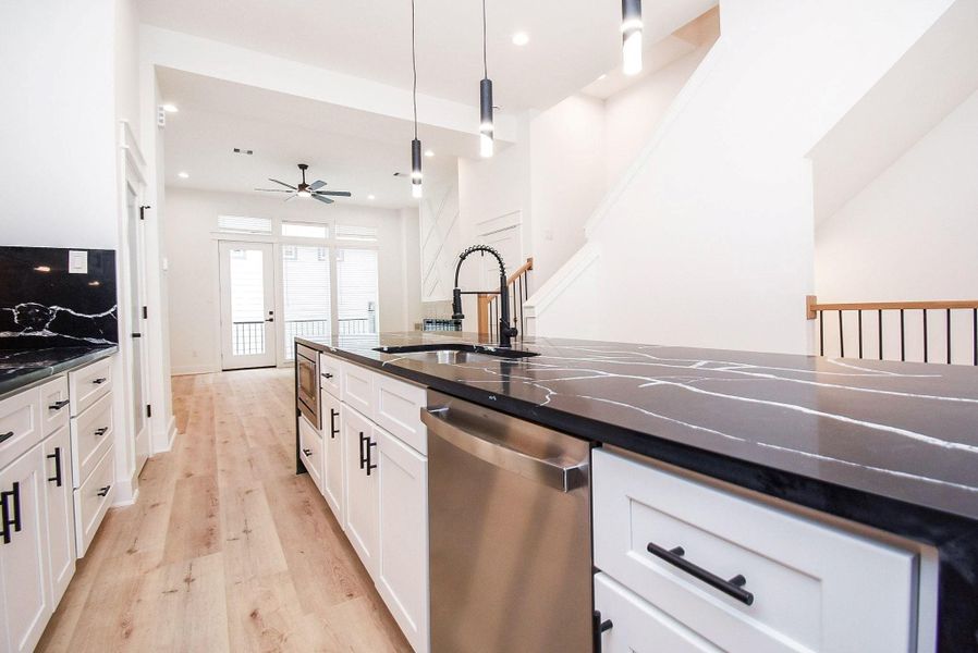 This modern kitchen features sleek black countertops with white cabinetry, a central island with a sink, and stainless steel appliances. The open layout flows into a bright living area with large windows and access to a balcony, complemented by light wood flooring and contemporary lighting.