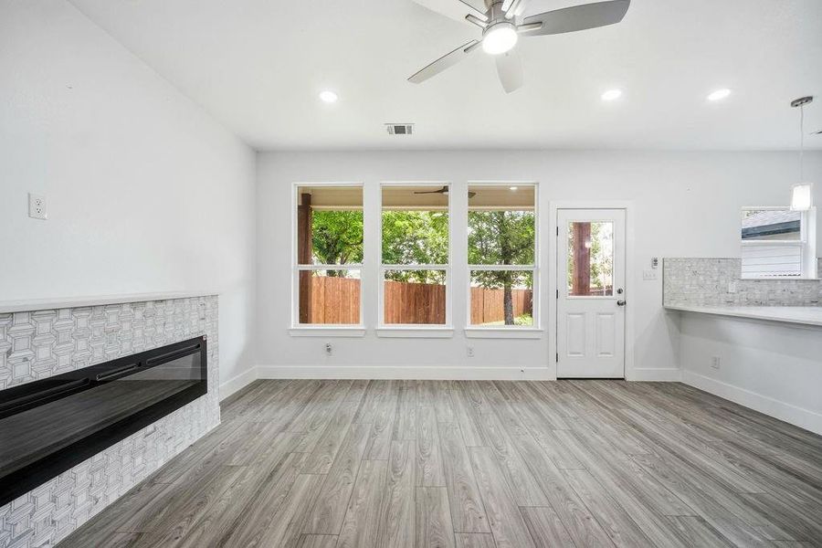 Spacious interior featuring wood-finish flooring, an electric fireplace with decorative tile surround, three large windows, recessed lighting, and a ceiling fan Spacious interior featuring wood-finish flooring, an electric fireplace with decorative tile surround, three large windows, recessed lighting, and a ceiling fan