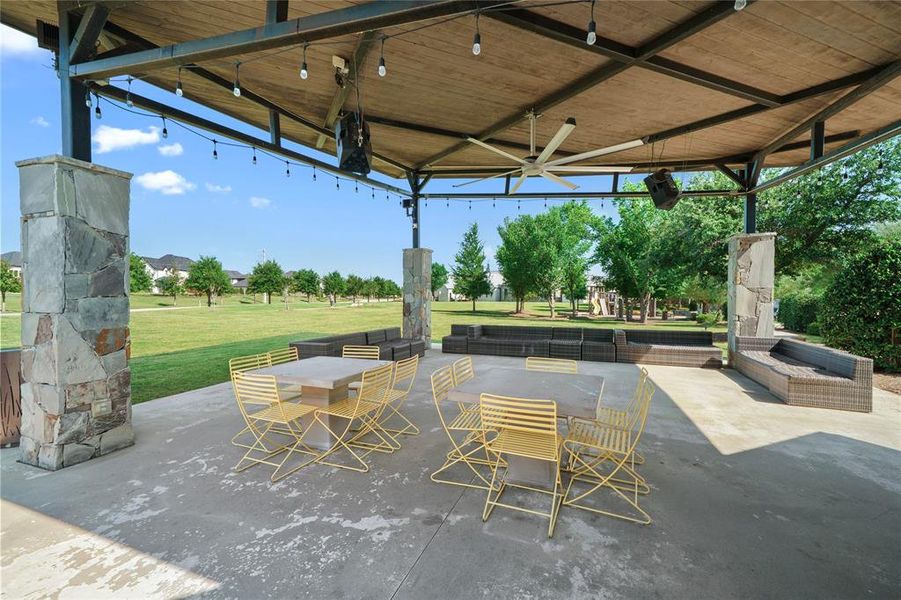 Covered patio area featuring stone-clad columns, a large ceiling fan, string lighting, and extensive concrete flooring