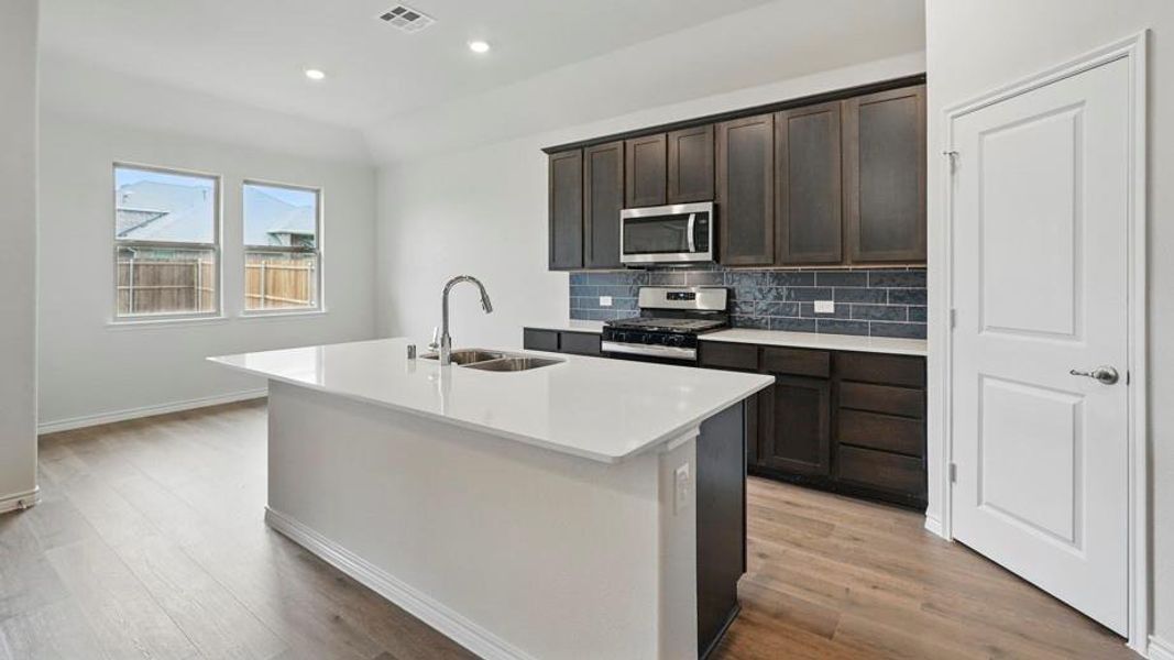 Spacious kitchen featuring a large island with an undermount sink and gooseneck faucet, dark wood-finish cabinetry, stainless steel appliances, and a subway tile backsplash