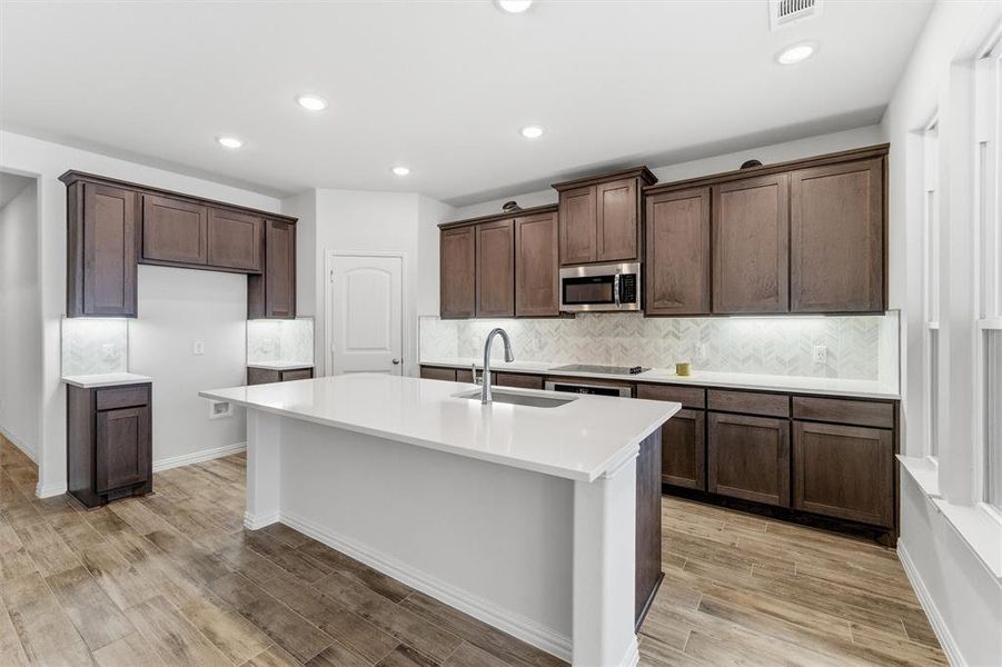 Kitchen featuring stainless steel microwave, light wood finished floors, a center island with sink, recessed lighting, and dark brown cabinets Kitchen featuring stainless steel microwave, light wood finished floors, a center island with sink, recessed lighting, and dark brown cabinets