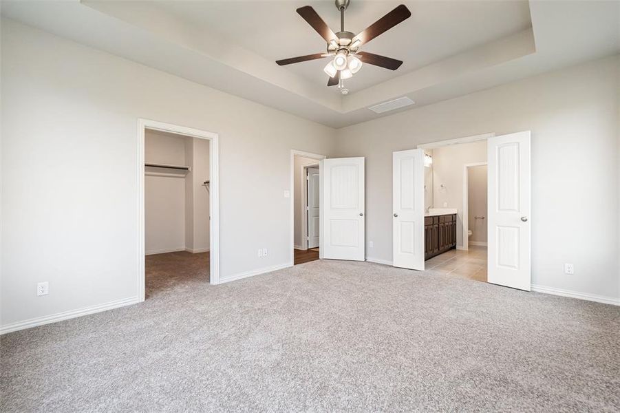 Unfurnished bedroom featuring a tray ceiling, a walk in closet, light colored carpet, ensuite bath, and ceiling fan
