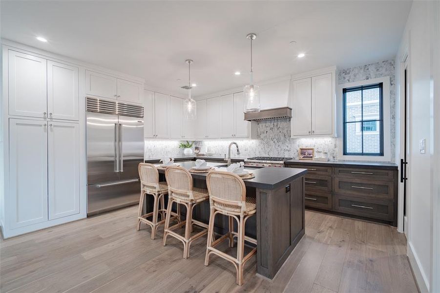 Kitchen featuring built in fridge, white cabinetry, hanging light fixtures, a kitchen island with sink, and custom range hood