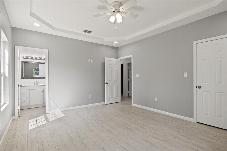 Unfurnished bedroom featuring a raised ceiling, light wood-type flooring, ceiling fan, recessed lighting, and ensuite bath