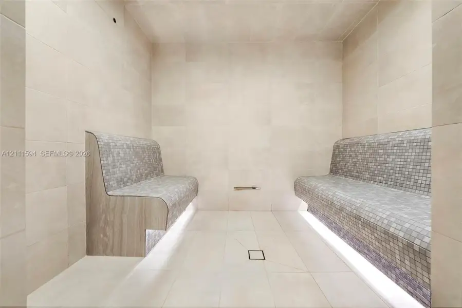 Steam room featuring large-format stone wall and floor tile, built-in dual benches with mosaic tile surfacing, integrated under-bench lighting, and a centered floor drain Steam room featuring large-format stone wall and floor tile, built-in dual benches with mosaic tile surfacing, integrated under-bench lighting, and a centered floor drain