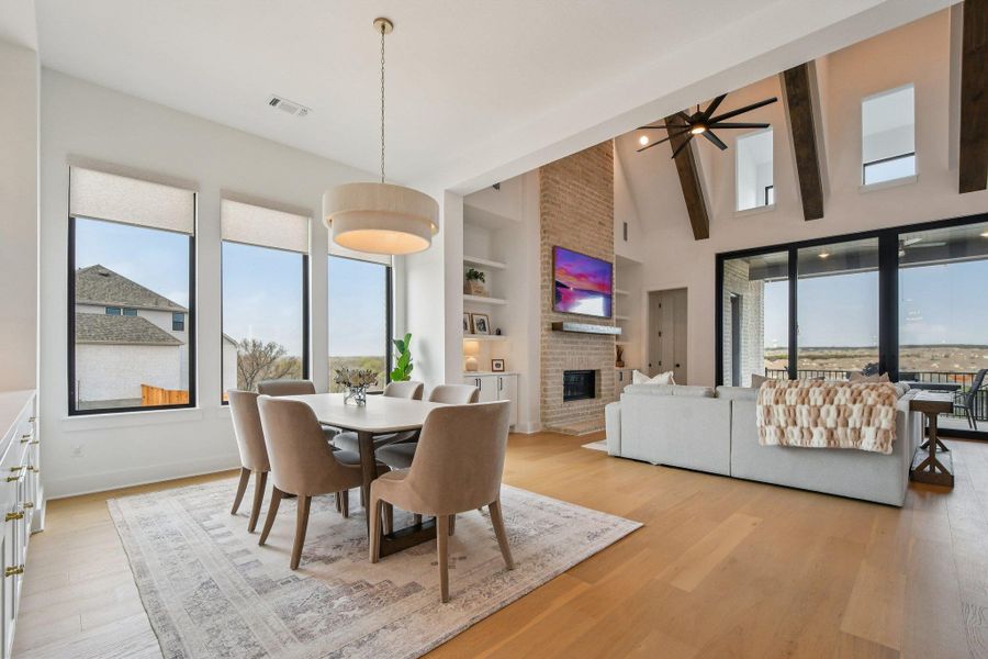 This open-concept living space features light wood flooring, a dining area with a modern chandelier, and a living area with a stone fireplace, high vaulted ceilings with exposed beams, and large windows and sliding glass doors that provide views of the outdoors This open-concept living space features light wood flooring, a dining area with a modern chandelier, and a living area with a stone fireplace, high vaulted ceilings with exposed beams, and large windows and sliding glass doors that provide views of the outdoors