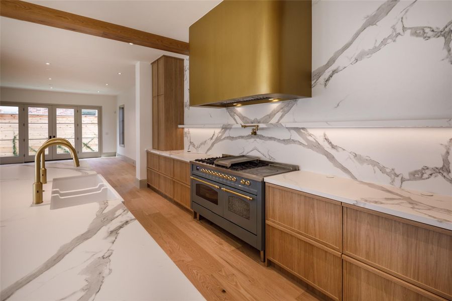 If you're going to make a hood the centerpiece, make it this one. A dramatic brass surround floats against book-matched veined stone, finished with warm wood cabinetry, integrated lighting, and brass details at the range.