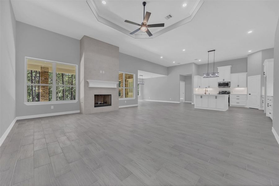 Sample of Home This spacious open-concept living area features a modern kitchen with white cabinetry and stainless steel appliances, a cozy fireplace, and large windows providing natural light. The neutral color palette and recessed lighting add a contemporary touch.