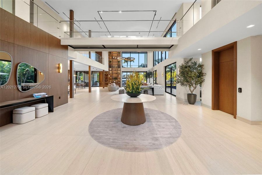 Expansive foyer with wide-plank light wood floors, walnut wall paneling, sculptural console with freeform mirror, marble round entry table, open second-floor glass balcony, and seamless flow into double-height great room with waterfront views.