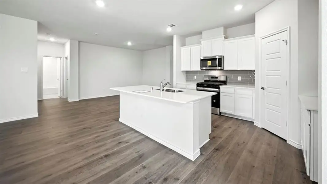Open-concept kitchen featuring an island with undermount sink, white cabinetry, dark subway tile backsplash, and wood-finish flooring Open-concept kitchen featuring an island with undermount sink, white cabinetry, dark subway tile backsplash, and wood-finish flooring
