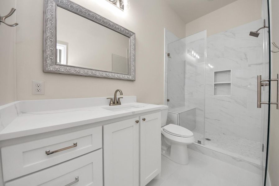 This en-suite bathroom features a modern design with a sleek white vanity, a large decorative mirror, and a spacious glass-enclosed shower with elegant tile work. The clean lines and neutral colors create a fresh and inviting atmosphere.