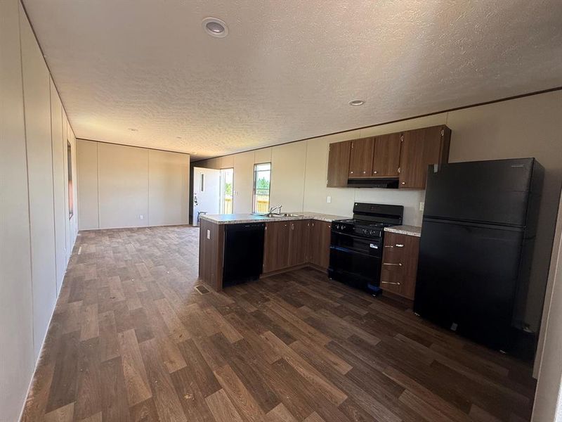 Open-concept kitchen featuring wood-finish cabinetry, black appliances including a refrigerator, stove, and dishwasher, and light-toned countertops Open-concept kitchen featuring wood-finish cabinetry, black appliances including a refrigerator, stove, and dishwasher, and light-toned countertops