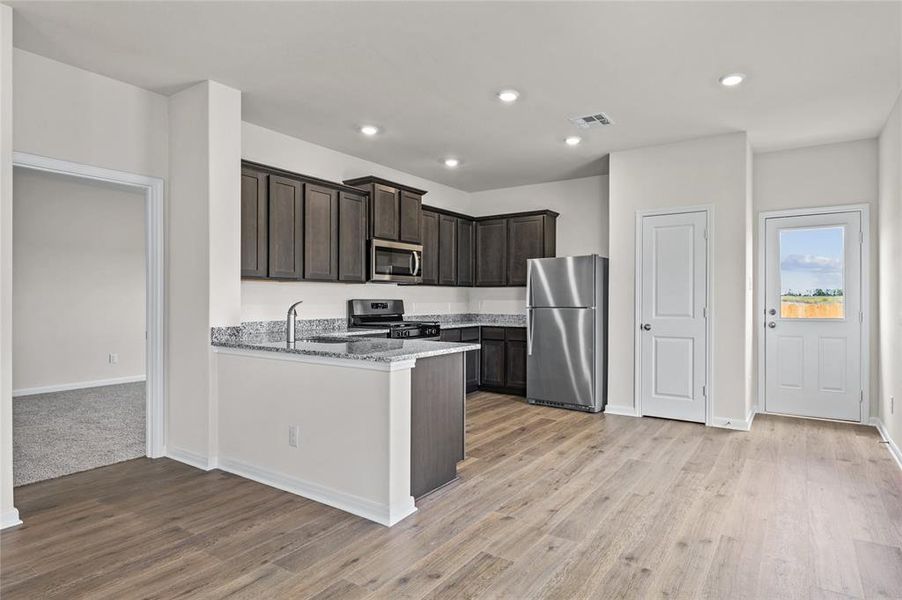 Kitchen featuring stainless steel appliances, dark brown cabinets, light stone counters, a peninsula, and light wood-style flooring