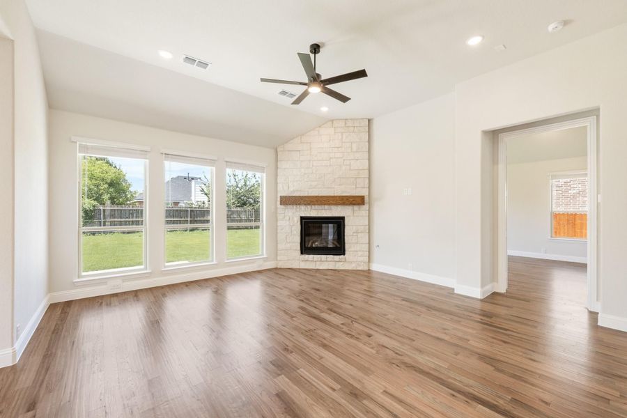 Open living room with hardwood floors, stone fireplace, ceiling fan, and large windows overlooking backyard Open living room with hardwood floors, stone fireplace, ceiling fan, and large windows overlooking backyard