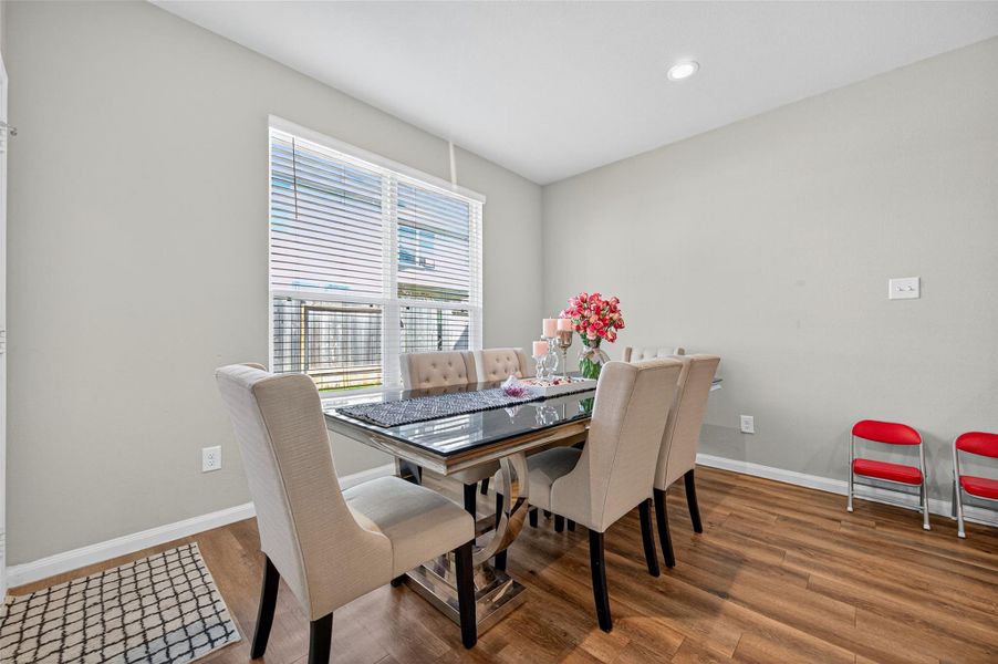 Bright dining area with modern decor, featuring a sleek table and comfortable upholstered chairs. Large window offers natural light, highlighting the wood flooring and neutral wall tones. Cozy and inviting space for dining.