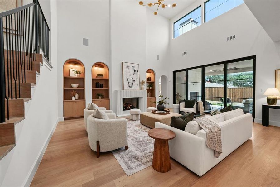 Living room featuring built in features, a fireplace with raised hearth, light wood-type flooring, stairs, and a towering ceiling Living room featuring built in features, a fireplace with raised hearth, light wood-type flooring, stairs, and a towering ceiling