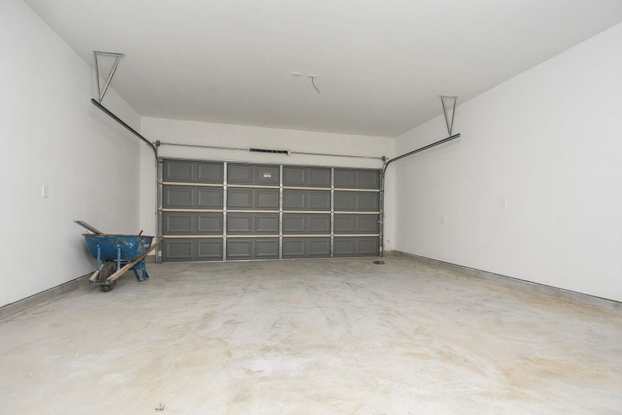 This is a spacious, clean two-car garage with a high ceiling, featuring a large metal door and automatic opener. There's a wheelbarrow present, indicating recent use or work. The floor is concrete and shows some stains, typical of garage use. This is a spacious, clean two-car garage with a high ceiling, featuring a large metal door and automatic opener. There's a wheelbarrow present, indicating recent use or work. The floor is concrete and shows some stains, typical of garage use.