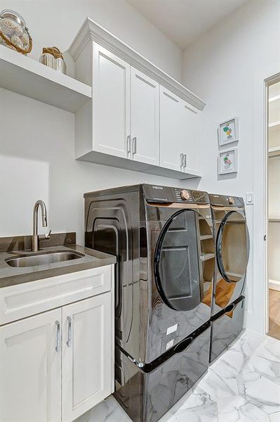 This downstairs laundry room adjoining the primary closet features sleek, modern appliances with a Samsung washer and dryer, a convenient utility sink, ample cabinetry for storage, and a clean, bright design with white finishes and tiled flooring.