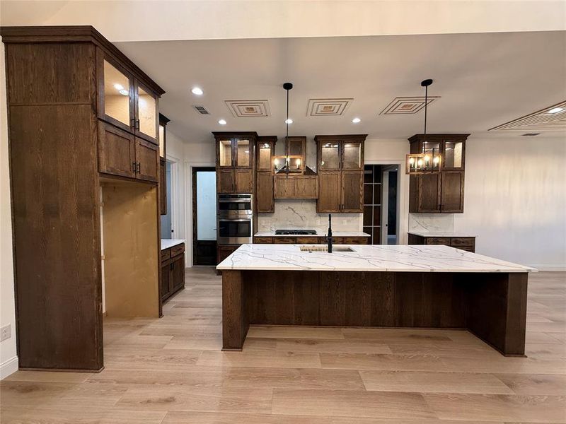 Kitchen featuring tasteful backsplash, a large island with sink, light stone counters, a breakfast bar area, and glass insert cabinets Kitchen featuring tasteful backsplash, a large island with sink, light stone counters, a breakfast bar area, and glass insert cabinets