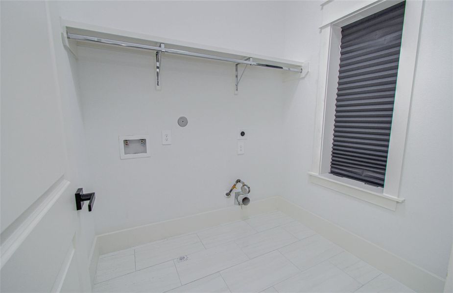 A bright, modern laundry room with hookups for a washer and dryer. It features white walls, tile flooring, a shelf for storage, and a window with a blind.