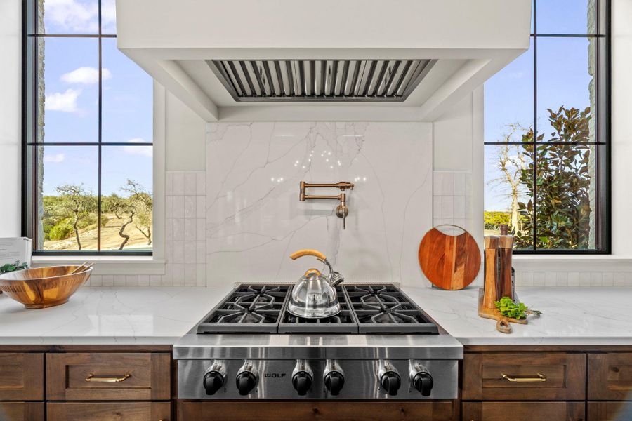 Kitchen with stunning windows that frame custom exhaust hood, stainless steel gas cooktop, light stone counters, and dark wood finish cabinets.