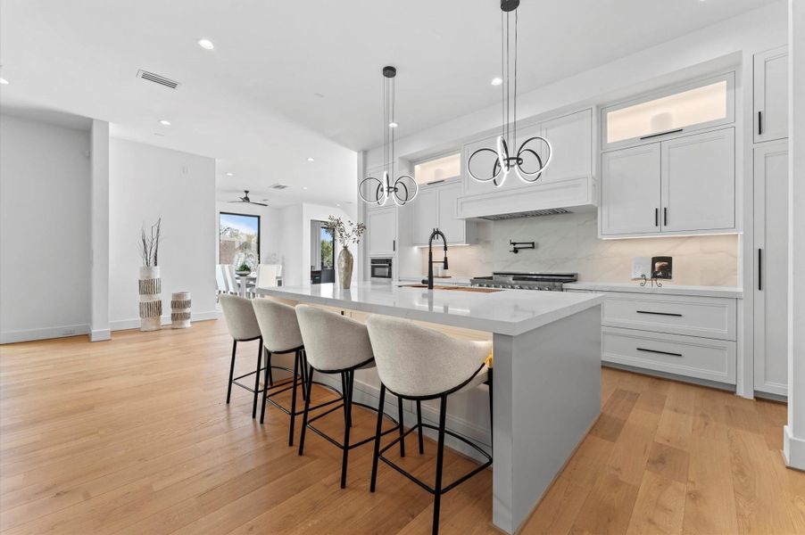 This modern kitchen features an open layout with a large island, sleek white cabinetry, and contemporary lighting fixtures. The hardwood floors add warmth, and ample natural light creates a bright and inviting space perfect for entertaining.