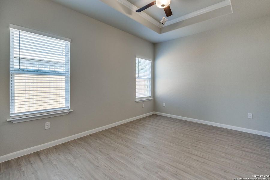 Spacious room featuring wood-finish flooring, two windows with blinds, a tray ceiling, and a ceiling fan with integrated lighting