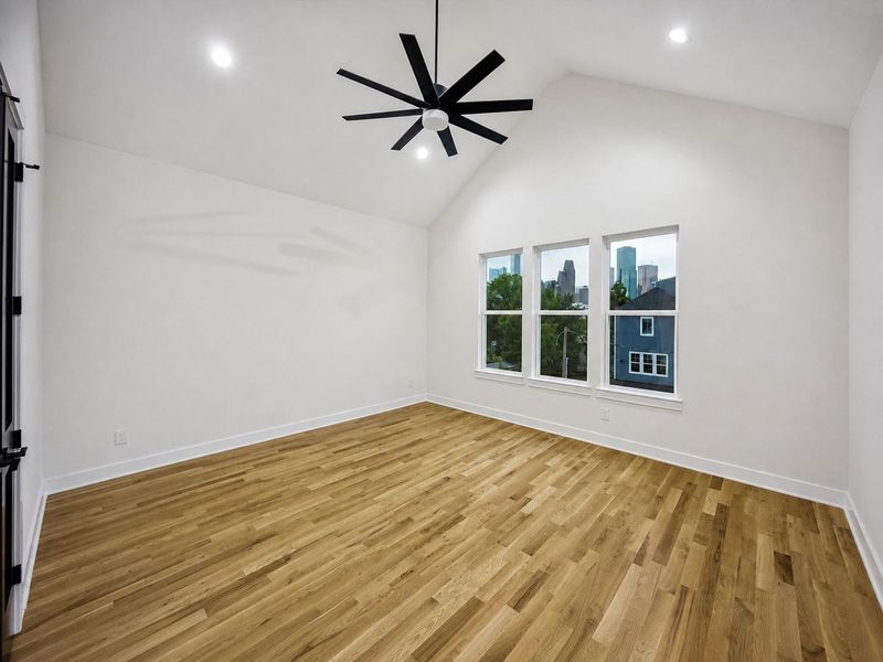 The expansive primary bedroom feels bright and impressive with a vaulted ceiling, warm wood flooring, recessed lighting, and a dramatic oversized black ceiling fan. Three tall windows frame attractive skyline views and fill the room with natural light. The generous proportions provide ample space for a full bedroom suite, seating area, or additional furnishings while maintaining an airy feel. The expansive primary bedroom feels bright and impressive with a vaulted ceiling, warm wood flooring, recessed lighting, and a dramatic oversized black ceiling fan. Three tall windows frame attractive skyline views and fill the room with natural light. The generous proportions provide ample space for a full bedroom suite, seating area, or additional furnishings while maintaining an airy feel.