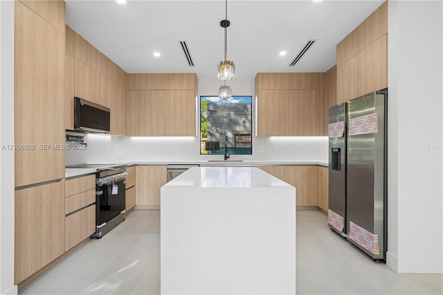 Modern kitchen featuring flat-panel wood-finish cabinetry, a large quartz-topped island, stainless steel appliances, under-cabinet lighting, and recessed ceiling lighting Modern kitchen featuring flat-panel wood-finish cabinetry, a large quartz-topped island, stainless steel appliances, under-cabinet lighting, and recessed ceiling lighting